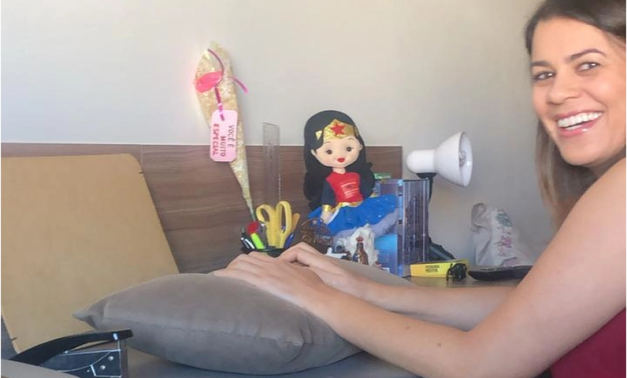 Mulher sorrindo sentada à mesa de trabalho em casa, com luminária, porta-lápis e bonecos na prateleira ao fundo