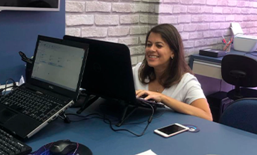 Mulher trabalhando sorrindo em frente a dois monitores no primeiro escritório externo da Co.Crie, parede de tijolo aparente ao fundo