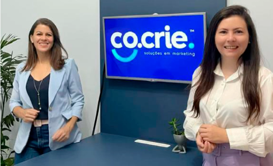 Duas sócias da Co.Crie em pé ao lado da mesa de reunião, com a logo da agência na TV atrás delas